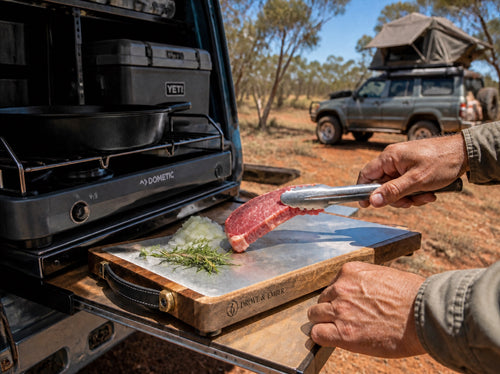 The Bedrock cutting board on a camp tailgate kitchen setup, steak being prepped with tongs, outback background