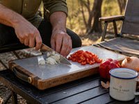 The Bedrock cutting board in use at a campsite, hands dicing vegetables with a camp mug and chair nearby